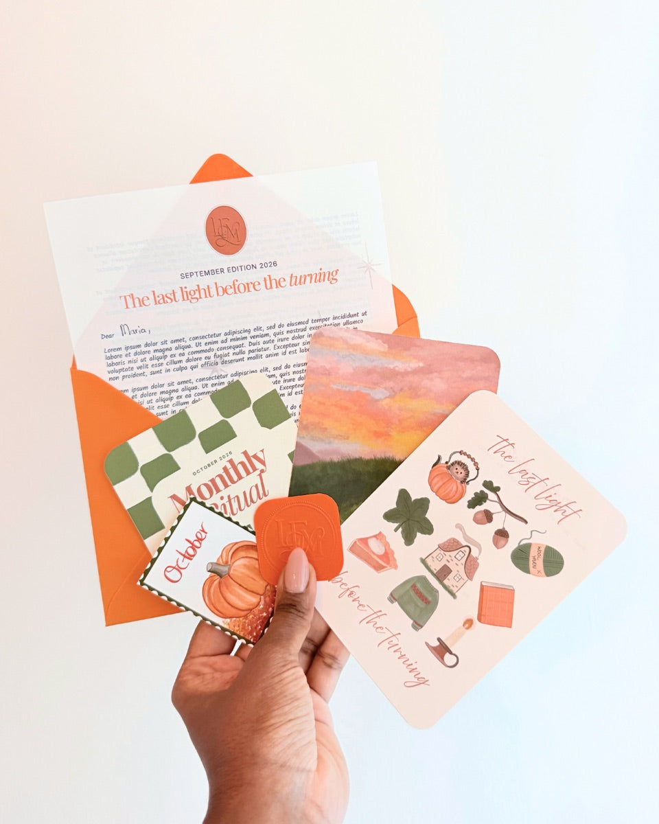 A hand holding the October Letters from Mashi edition — the orange envelope, autumn letter, pumpkin stamp, sunset art print, and Monthly Ritual card